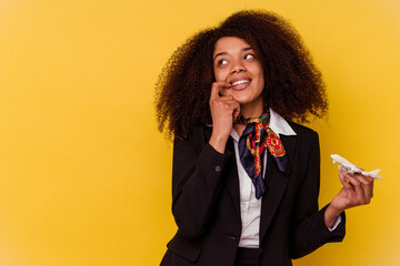 Young african american air hostess holding a little plane isolated on yellow background relaxed...