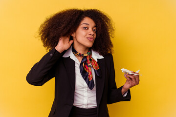 Young african american air hostess holding a little plane isolated on yellow background trying to...