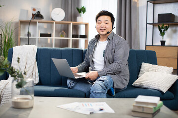 Handsome asian man using headset and laptop for video conversation. Male freelancer in casual wear sitting on couch and having online meeting.