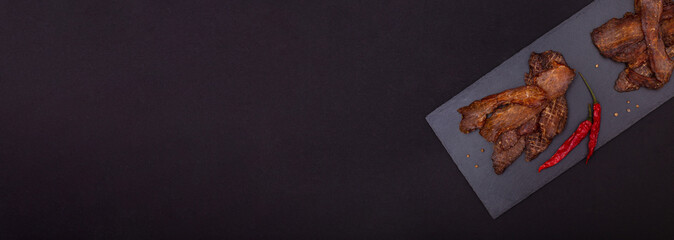 Banner with jerky meat snacks, red papper and peppercorns on black stone cutting board on dark...