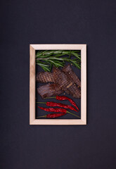 Jerky snacks, red papper and rosemary inside of wooden frame on black background. Creative concept with dried spiced meat for beer
