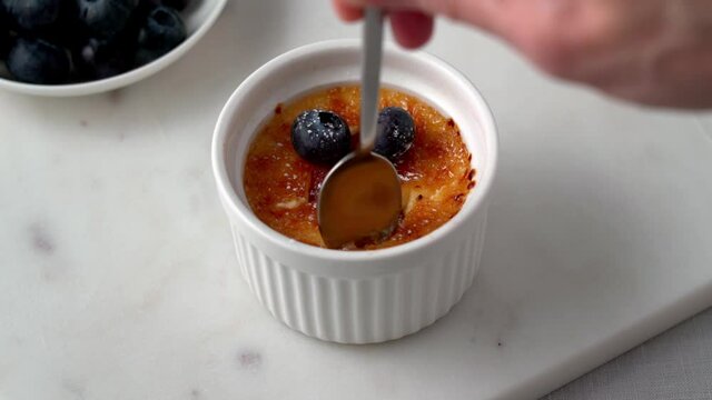 Spoon breaking crispy caramelized crust into creme brulee in ramekins. Eating sweet french dessert or spanish creme catalana. Burned Trinity or Cambridge burnt cream. 