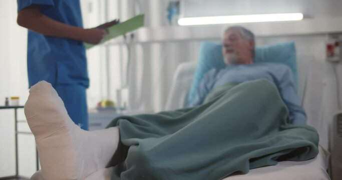 Doctor talking to senior man patient with broken leg lying in hospital bed