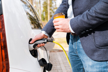 Close up man inserting power cable supply for charging electric car and holding cup of coffee. 
