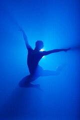 Ballerina in black bodysuit dancing with blue light and smoke. Silhouette of dancer in ballet shoes.
