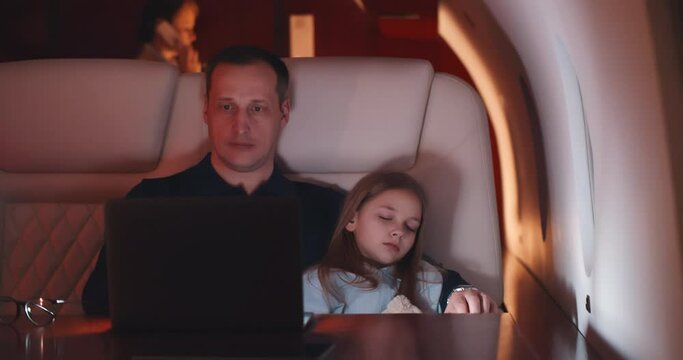 Businessman working on laptop and holding sleeping daughter flying on private jet at night