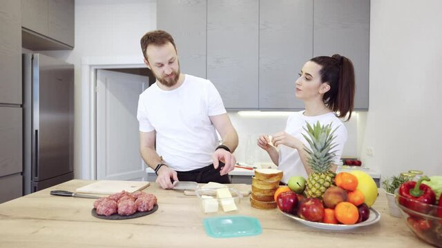 Young cheerful couple cooking food together. They are preparing meat burgers at home in the kitchen. Man slices cheese, and woman wants feeds him