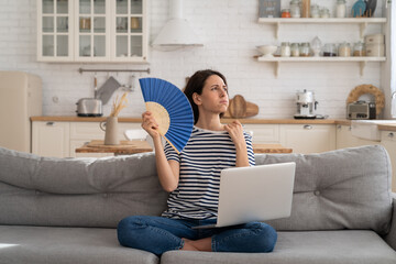 Tired millennial woman suffering from heatstroke flat without air-conditioner, waving blue fan,...