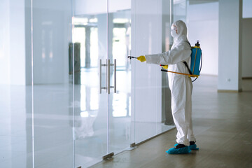 Man in Hazmat suit, protective gloves and goggles use sprayer equipment disinfect  in office. Protection agsinst COVID-19 disease. - obrazy, fototapety, plakaty