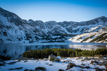 Sunny day in Tatra Mountain valley, Poland. Fresh snow, small dwarf mountain pines and a lake which is starting to freeze. Selective focus on the ridge, blurred background.