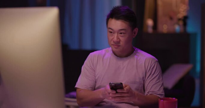 Chinese handsome relaxed young man iwth mobile phone using pc computer workingon freelance project and checking time. Home interior.