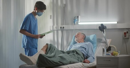 Surgeon in medical mask examining aged male patient with broken leg