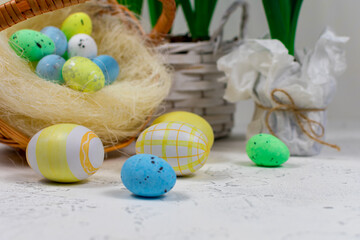 Easter eggs in pastel colors on a white table against the background of a basket with Easter eggs and flowers of daffodils. Selective focus. Place for an inscription. Close-up.
