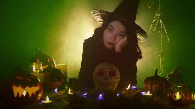 Bored witch sits surrounded by jack o'lanterns and skull. Young caucasian woman in dark cloak and a hat is sighing among Halloween decorations. Celebrating a holiday in a mystical voodoo installation.