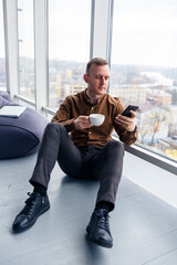 Handsome young male manager is sitting on the floor with the phone near the panoramic window. Male businessman working on a new project