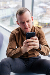 Handsome young male manager is sitting on the floor with the phone near the panoramic window. Male businessman working on a new project