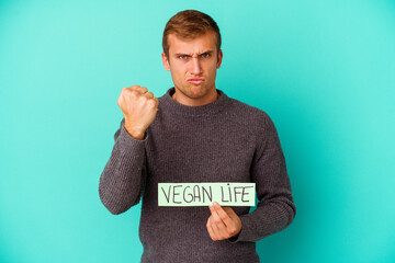 Young caucasian man holding a vegan life placard isolated on blue background  showing fist to camera, aggressive facial expression.