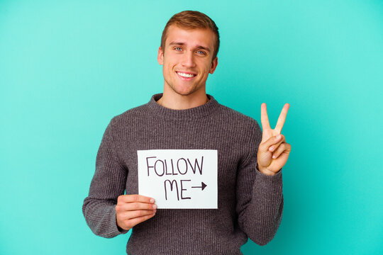 Young caucasian man holding a Follow me placard isolated on blue background showing number two with fingers.