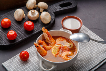 Tom yam soup with shrimps in a tureen with a spoon on a concrete background next to a bowl with coconut milk mushrooms and tomatoes.