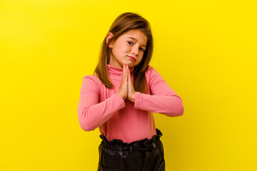 Little caucasian girl isolated on yellow background holding hands in pray near mouth, feels...