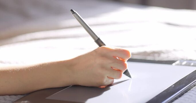 Close up view of woman hand using digital tablet drawing with stylus pen. Designer working on bed at home.