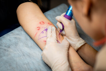 Master tattoo draws the red paint on the clients tattoo. Tattoo artist holding a pink tattoo machine in black sterile gloves and working on the professional blue mat.