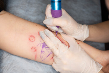 Master tattoo draws the red paint on the clients tattoo. Tattoo artist holding a pink tattoo machine in black sterile gloves and working on the professional blue mat.