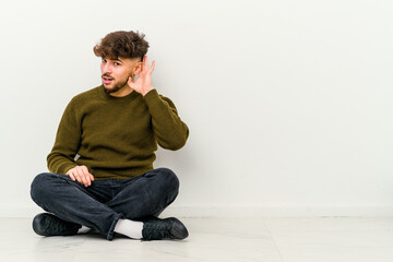 Young Moroccan man sitting on the floor isolated on white background trying to listening a gossip.