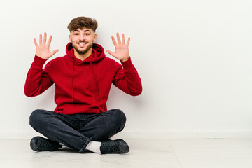 Young Moroccan man sitting on the floor isolated on white background showing number ten with hands.