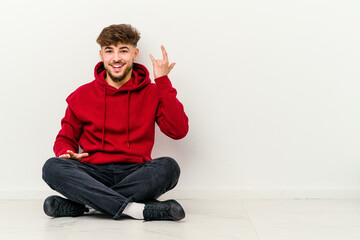 Young Moroccan man sitting on the floor isolated on white background showing rock gesture with fingers