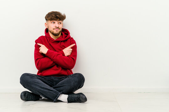Young Moroccan man sitting on the floor isolated on white background points sideways, is trying to choose between two options.