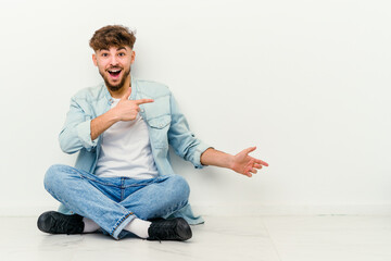 Young Moroccan man sitting on the floor isolated on white background excited pointing with forefingers away.