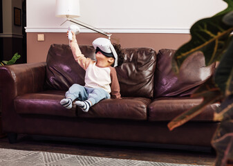 Little boy in comfy clothes using VR set while sitting on a sofa. Kid raising hand with a joystick.