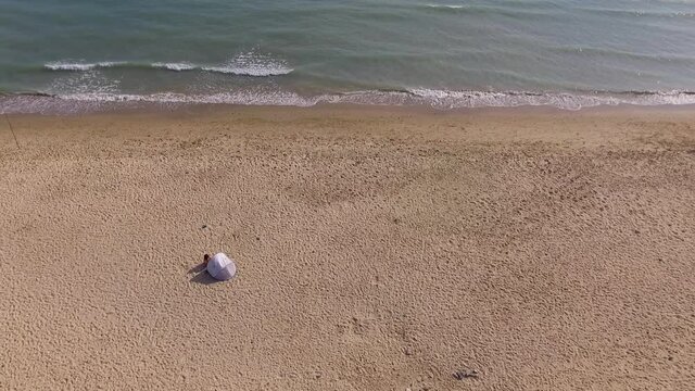 Aerial view of the beach paralell to the sea. Small waves ondulate the sea shore right next to the sand. Only a small tent with someone next to it and also another  small group of people are seen.
