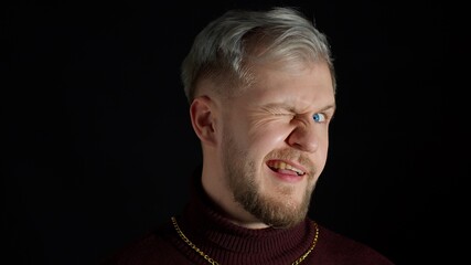 Stylish smiling young man with blue eyes in trendy clothes looking at camera, charming smile, winking, blinking eye, flirting, feeling happiness, fun and enjoyment on black background. People emotions