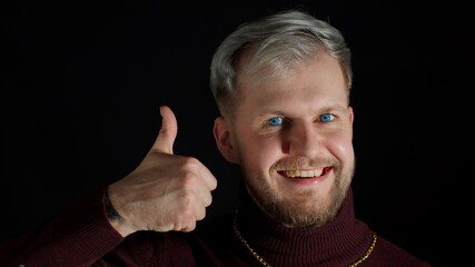 Cheerful smiling man in stylish blouse looking approvingly at camera, nods his head, showing thumbs up on black background. Facial expression of bearded positive guy with gray hair. Modern fashion