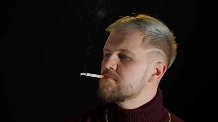Fototapeta premium Stylish addicted young man with blue eyes in trendy clothes smoking cigarette, looking at camera, feeling happiness, fun and enjoyment on black studio background. People bad habits. Modern fashion
