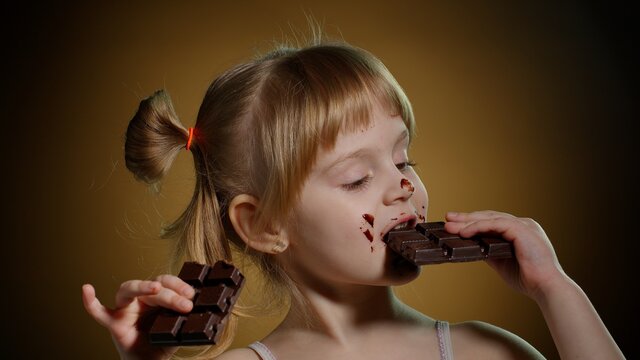 Happy Smiling Little Girl Eating Milk Chocolate Bar Isolated On Dark Background. Satisfied Face Of Pretty Caucasian Child Enjoying Unhealthy Sweet Food Indoors. Joyful Kid Eating Dessert Sweet Candies