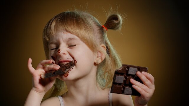 Happy Smiling Little Girl Eating Milk Chocolate Bar Isolated On Dark Background. Satisfied Face Of Pretty Caucasian Child Enjoying Unhealthy Sweet Food Indoors. Joyful Kid Eating Dessert Sweet Candies