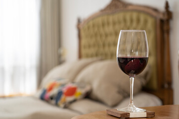 Glass of red wine on a table, behind a bed with a stylish back, and window. Day time. Room place.