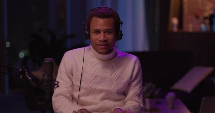 Black handsome young casual man in white sweater recording electronic music audiotrack at home. Male portrait. Musician performance. Microphone.