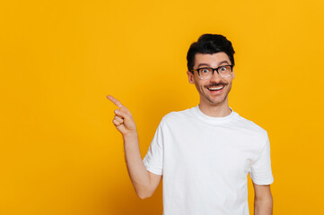 Excited caucasian happy guy in white basic t-shirt and glasses amazed looks at camera and points finger to the side at space for your presentation, stands on isolated orange color background, smiling
