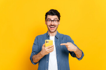 Happy joyful caucasian stylish guy with glasses, holding smartphone in hand and pointing at it...