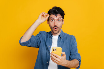 Amazed handsome unshaven caucasian guy holding smartphone in his hand, looking at the camera in...
