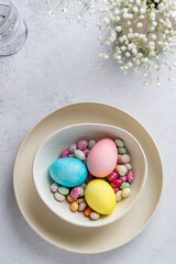 Fototapeta premium Festive Easter table with small white flowers, colored eggs and colorful speckled chocolate candies on ceramic plate. Top view