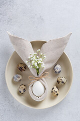 Easter table decoration with an egg in napkin in the shape of an Easter bunny with ears, with small white flowers and quail eggs on ceramic plate. Top view, copy spase.