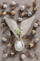 Stylish Easter background with egg in napkin in the form of an Easter bunny with ears, with small white flowers, quail eggs and speckled feathers on sacking. Top view.