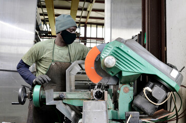 Black worker with protective mask for Covid-19 virus cuts a piece of material with a circular saw machine. Construction of a homemade muffler for sportive cars in stainless steel or industrial use.