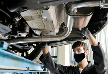 Young Mechanic With Protective Mask for Covid-19 virus on the Mouth Installing Exhaust Pipe Under a...