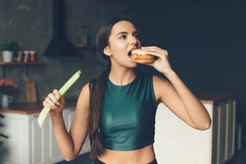 Brunette sporty woman is eating a burger while holding a leek in her hands
